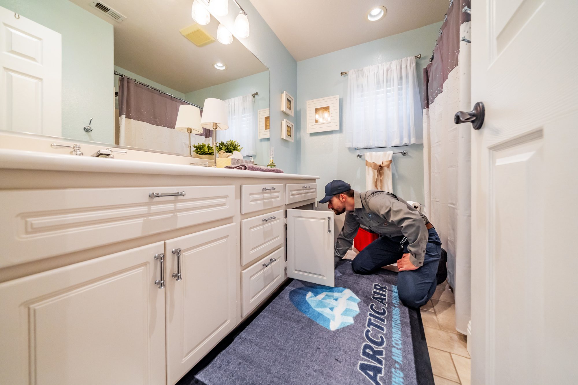 Arctic Air plumber kneeling on a branded mat to look inside an open bathroom vanity cabinet