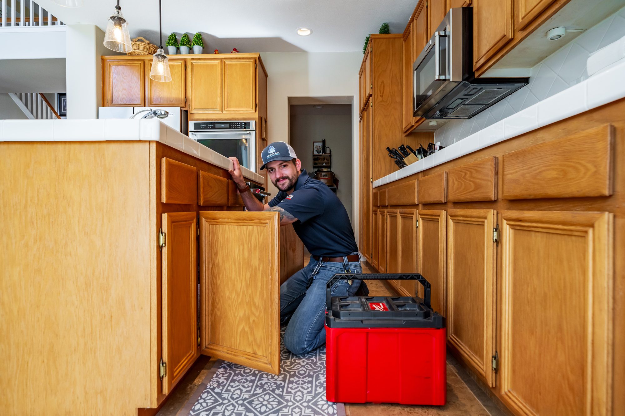 Arctic Air plumber kneeling at an open kitchen sink cabinet beside a red tool case